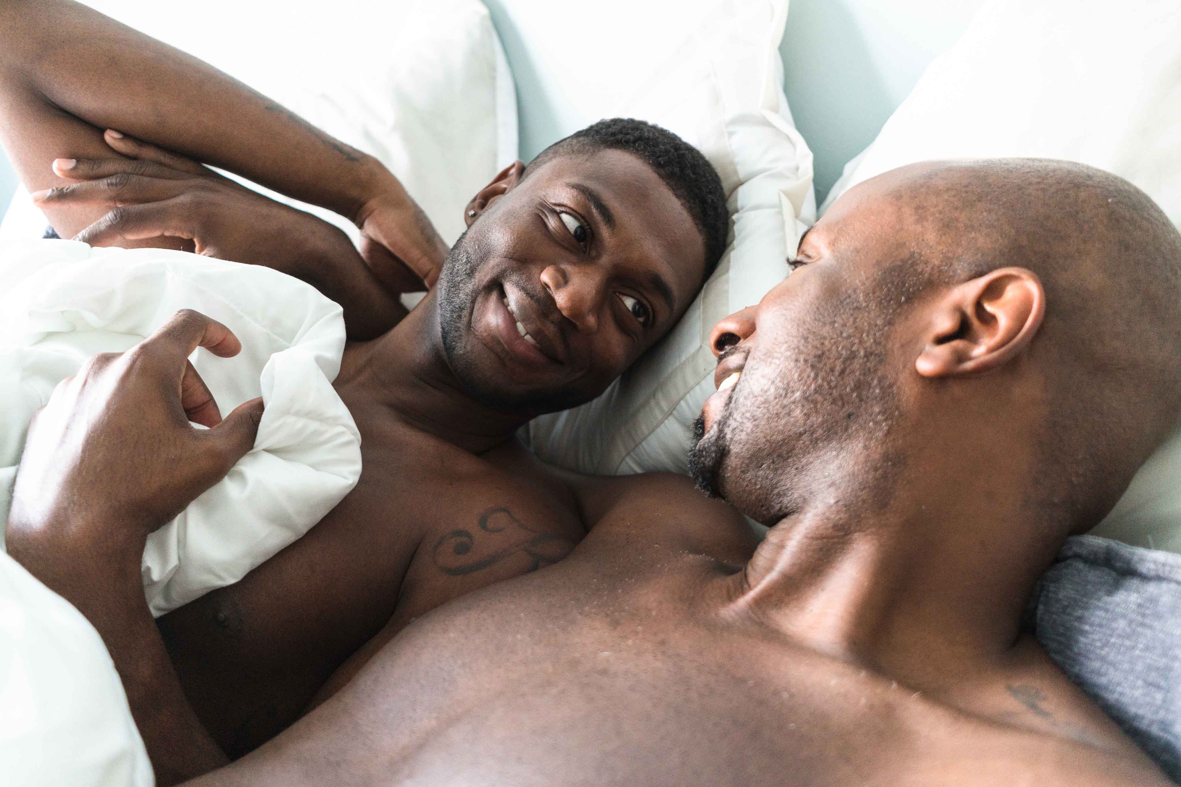 Two dark-skinned men laying in bed with each other.