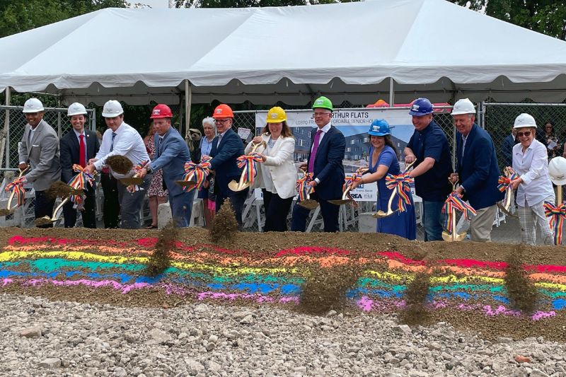 A group of diverse women and men wearing colorful hardhats and holding gold shovels with rainbow ribbons. They are throwing dirt from a dirt hill decorated with rainbow chalk onto a pile of gray rocks in front of them.