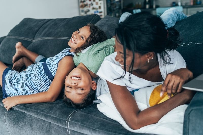 A dark-skinned woman and two medium-skin kids laying on top of each other on a couch smiling.