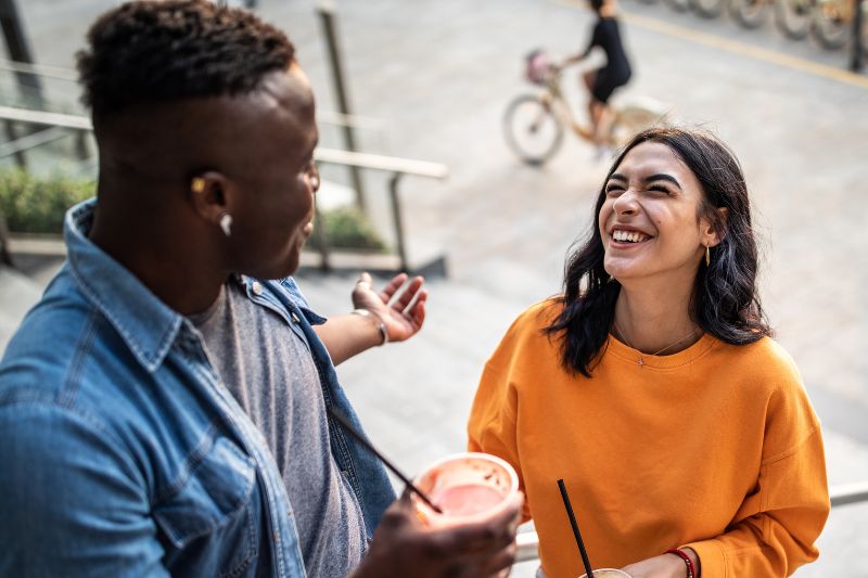 A dark-skinned man and a medium-skinned woman talking and laughing while drinking smoothies on a side walk.