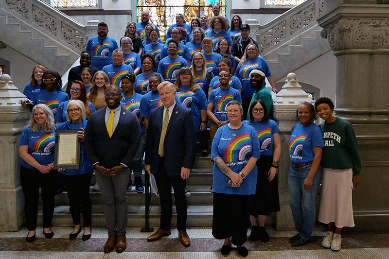 Caracole Staff wearing matching blue progressive pride shirts standing on the staircase of Cincinnati City Hall. Caracole CEO, Linda Seiter, a light-skinned woman wearing glasses, is holding Caracole's Resolution of Honor. She stands by City Council members, Reggie Harris and Mark Jeffreys.