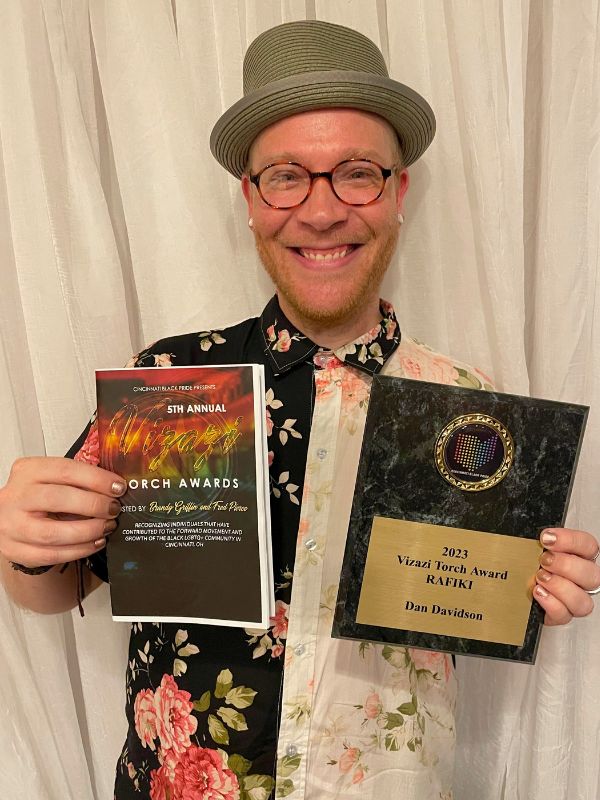 Dan Davidson, a light-skinned man holding his 2023 Rafiki Vizazi Torch Award and a program for the 5th Annual Vizazi Torch Awards.