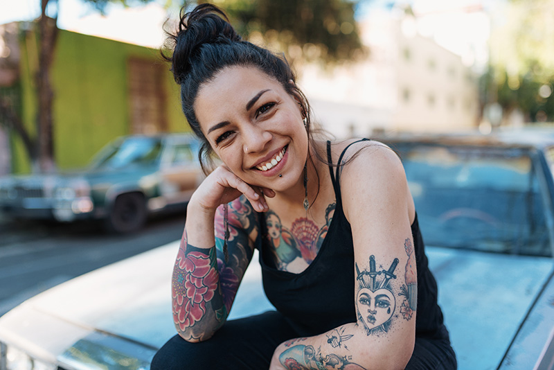 A smiling, medium skin toned woman with tattoos sits on car hood