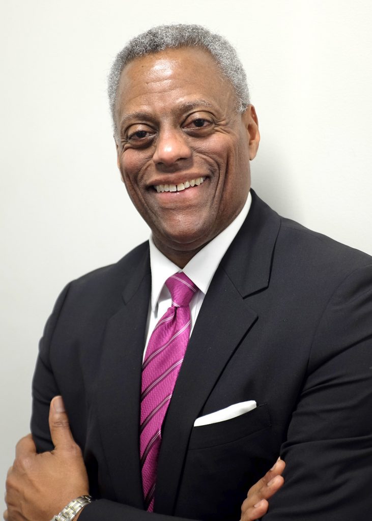 Jesse Milan Jr., JD, a dark-skinned man wearing a dark navy suit and a fuchsia tie with silvery stripes, smiling in front of a blurred gray background. He is President & CEO of AIDS United until his retirement on July 1, 2025.