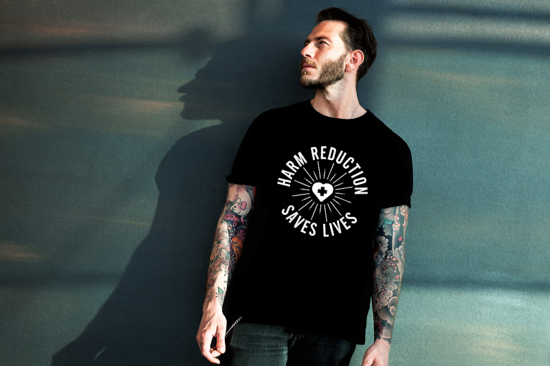 Light-skinned man leaning against gray wall wearing a black t-shirts that reads, "Harm reductions saves lives."
